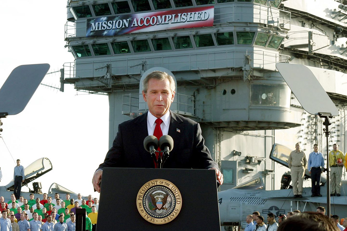 George W. Bush stands at a podium on an aircraft carrier in front of a banner that reads "Mission Accomplished"
