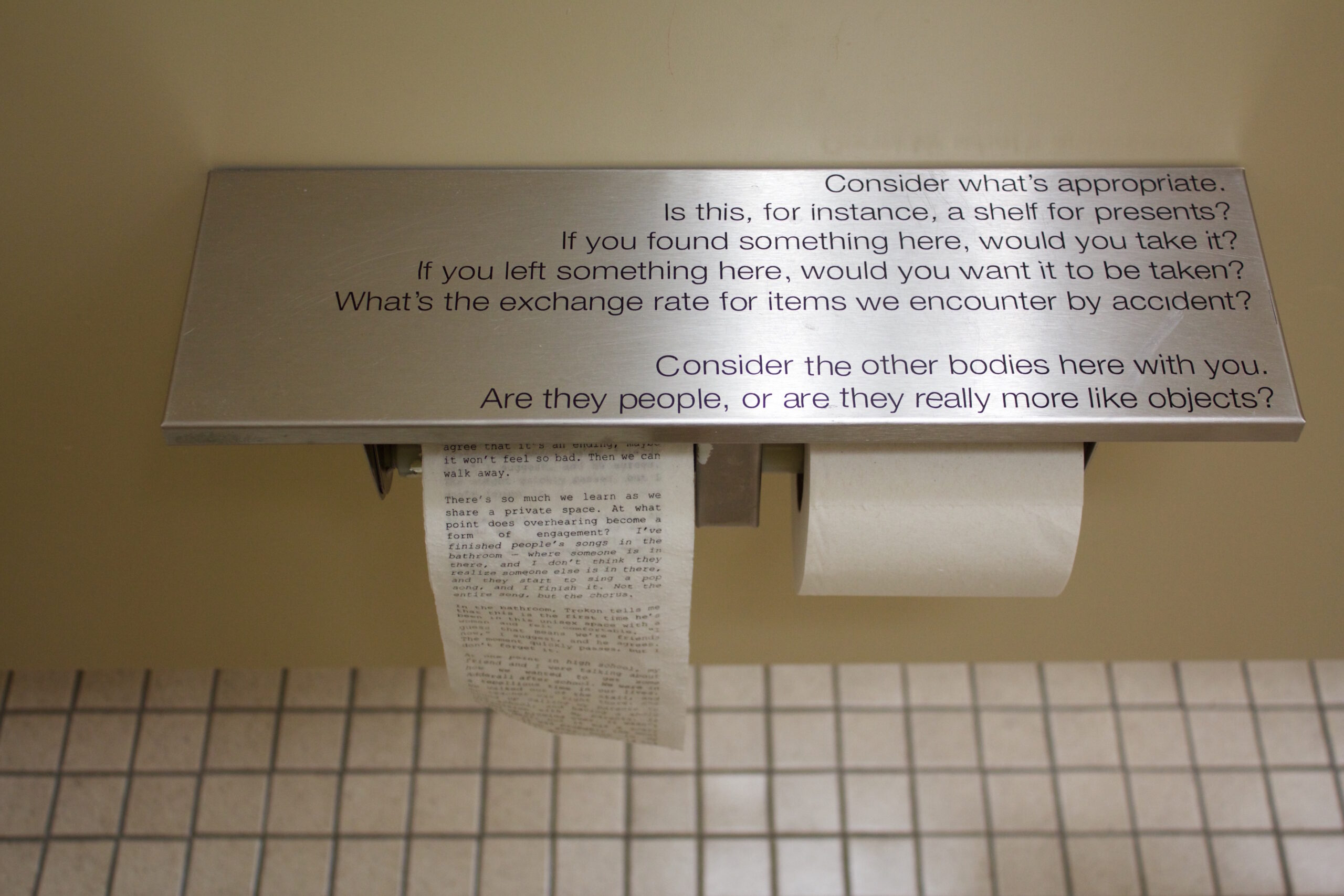 A stainless steel shelf in a bathroom stall holds two rolls of toilet paper, one printed with words. The shelf asks questions about how the reader feels about the bathroom.