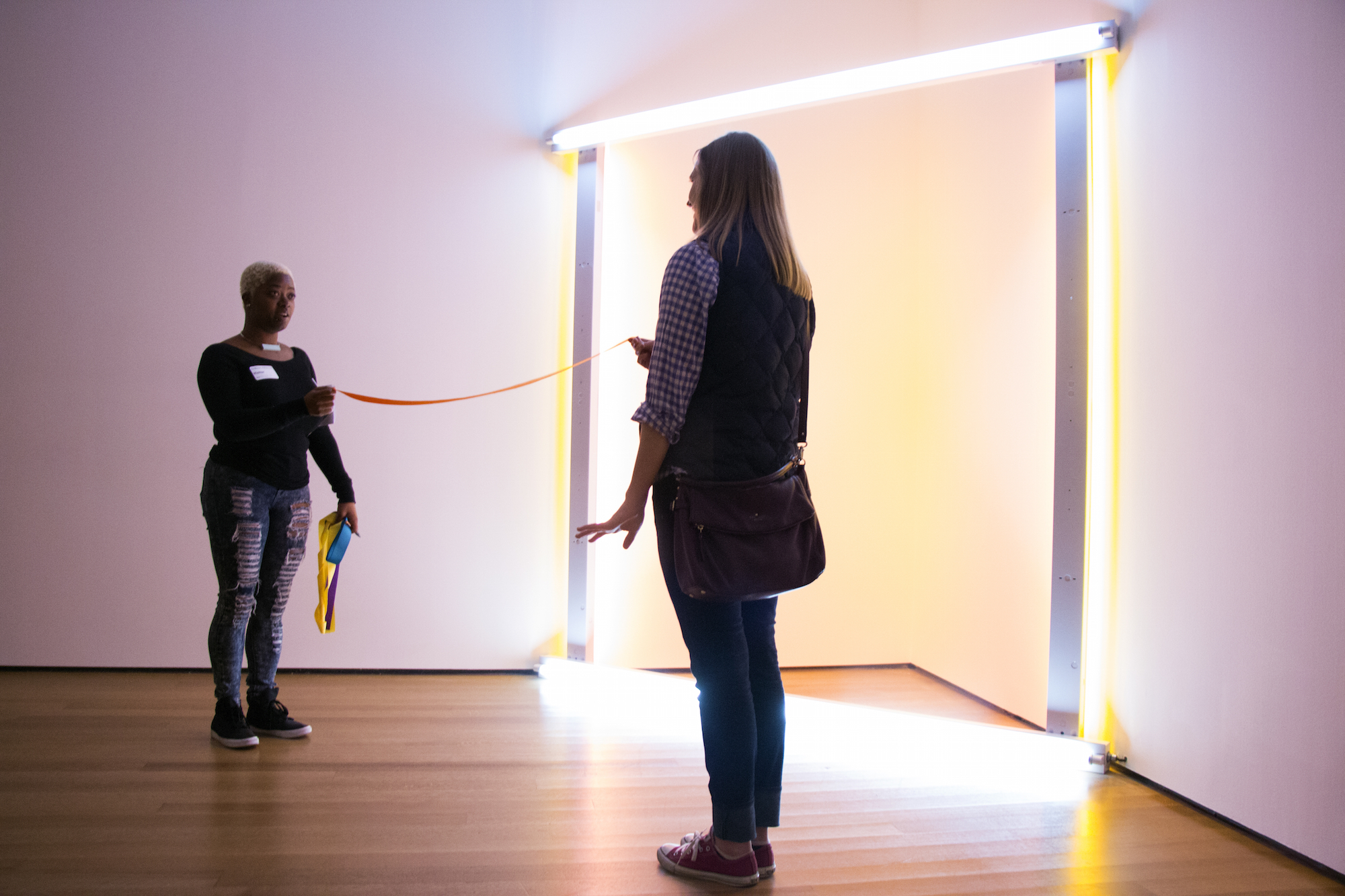 Two people stand in front of a light sculpture holding the ends of a tape measure that stretches between them.
