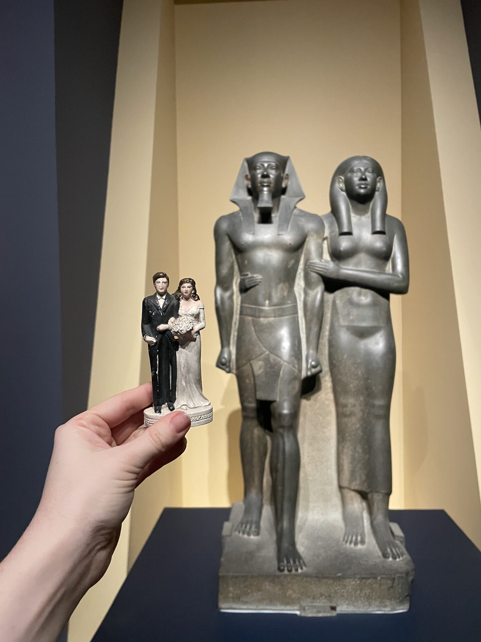 A white hand holds a bride and groom wedding topper in front of a large Egyptian statue couple