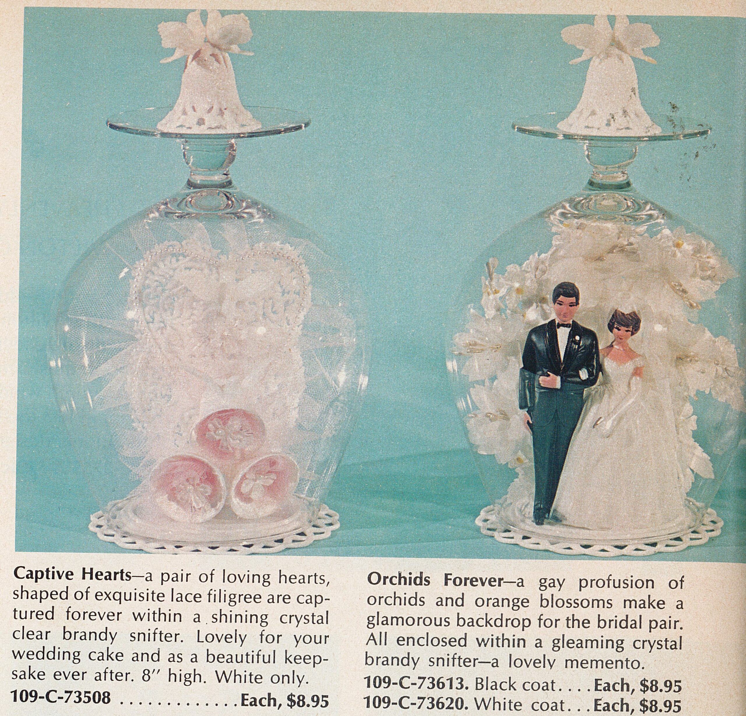 Two cake toppers, a set of bells with a heart and a bride and groom couple, sit on top of doilies and inside brandy snifter glasses