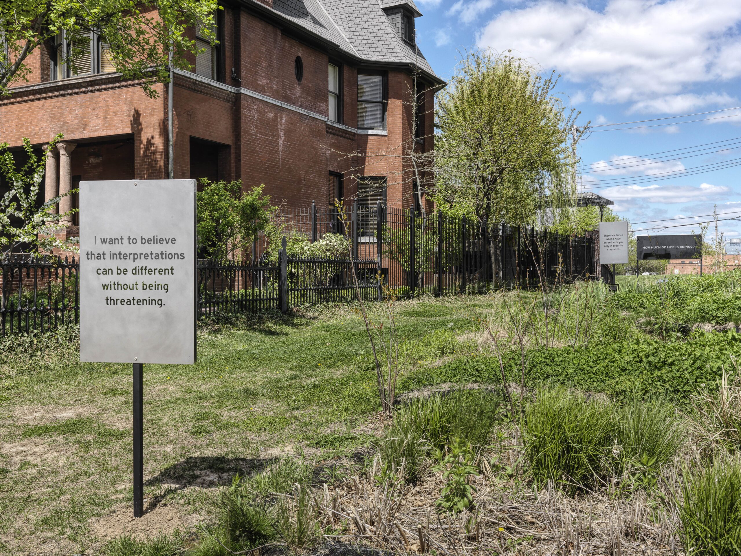 A metal sign in a field in front of a brick house reads "I want to believe that interpretations can be different without being threatening."