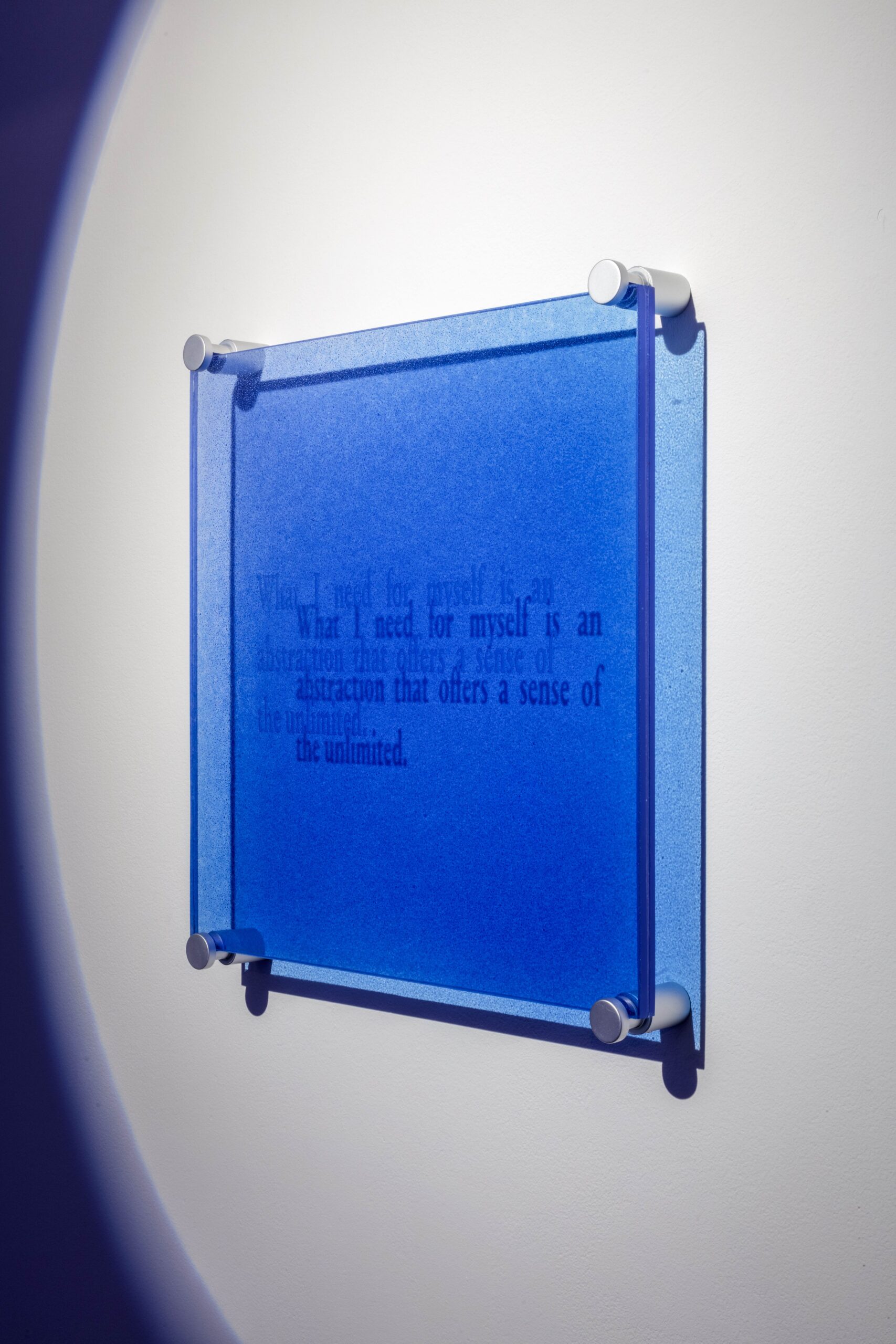 A blue glass square mounted on a wall, seen sideways, reads "What I need for myself is an abstraction that offers a sense of the unlimited."